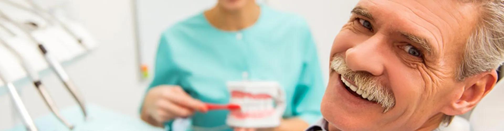 An older man smiling while a blurred dentist in the background holds and brushes a tooth model.