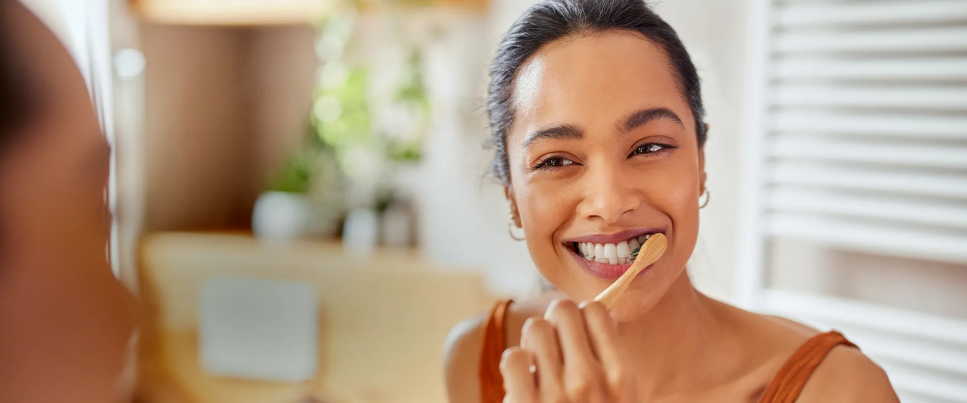 A cheerful young woman brushes her teeth using a bamboo toothbrush while smiling at her reflection in the bathroom mirror. The setting is bright and warmly lit, with indoor plants adding a refreshing touch to the space. This image conveys a focus on sustainable dental practices and personal care.