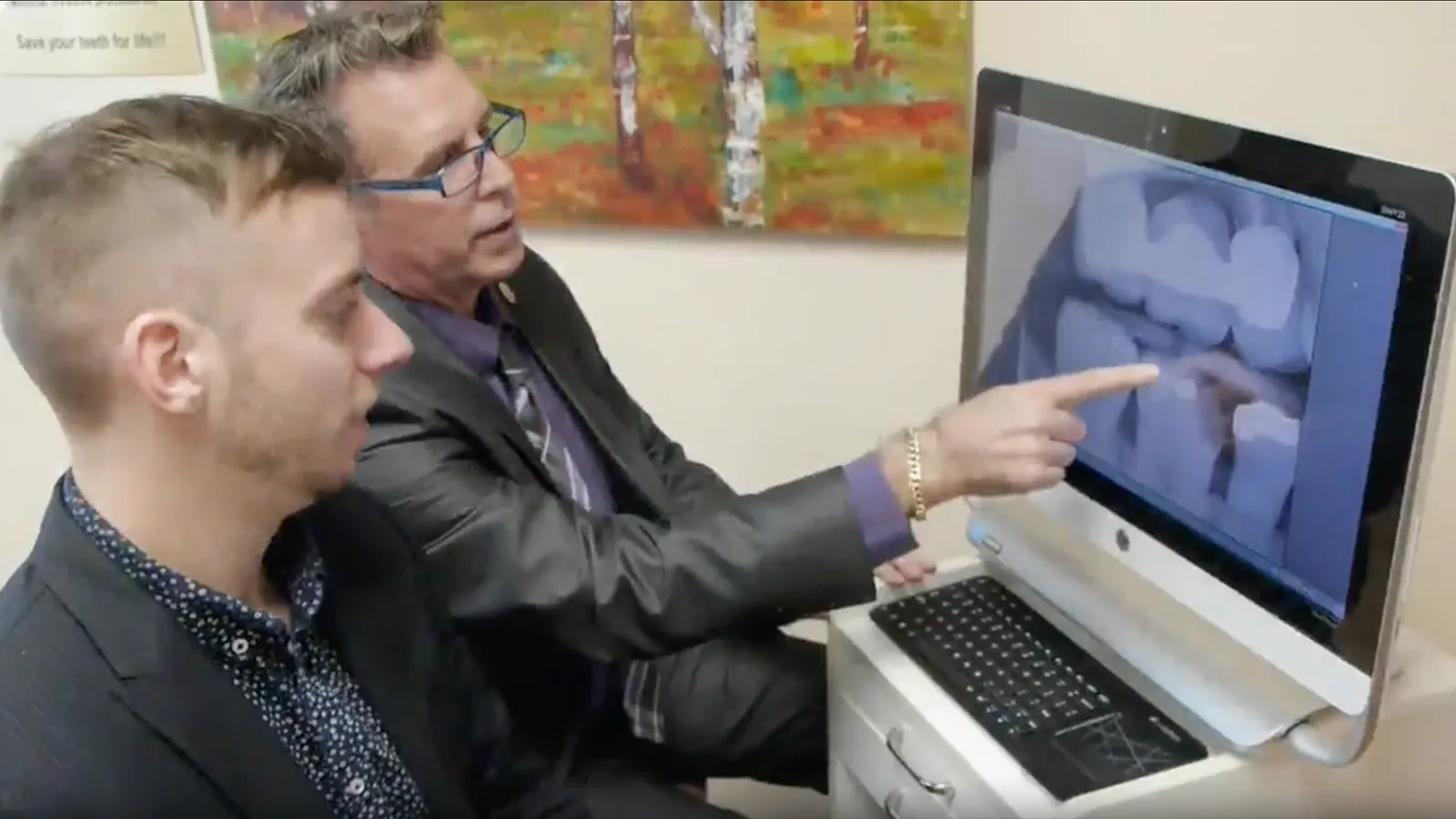 Thumbnail of a doctor pointing to dental X-rays on screen while seated beside patient.