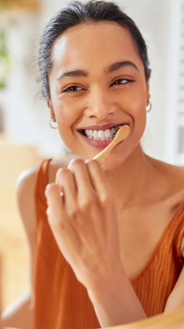 A cheerful young woman brushes her teeth using a bamboo toothbrush while smiling at her reflection in the bathroom mirror. The setting is bright and warmly lit, with indoor plants adding a refreshing touch to the space. This image conveys a focus on sustainable dental practices and personal care.